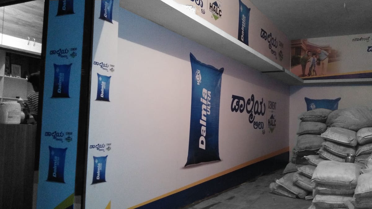 Cement shop interior with printed Dalmia Ultra wall graphics in Kannada beside stacked cement sacks.