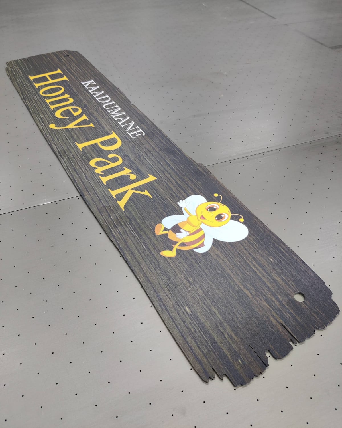 Contour-cut board printed with wood grain and the words Kaadumane Honey Park, lying on a perforated flatbed printer bed.