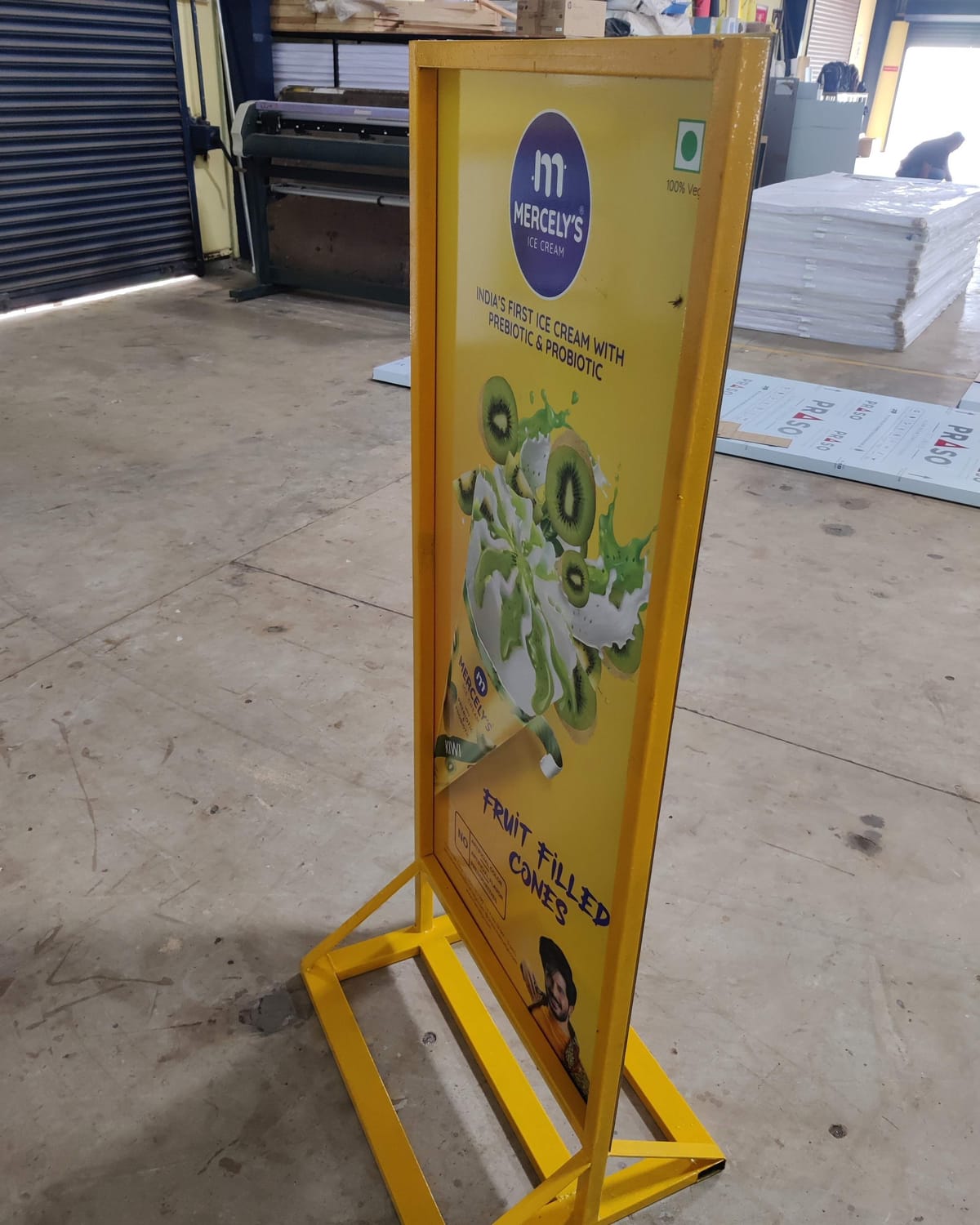 Yellow steel-framed floor standee printed with Mercely's ice cream artwork, standing on a workshop floor.