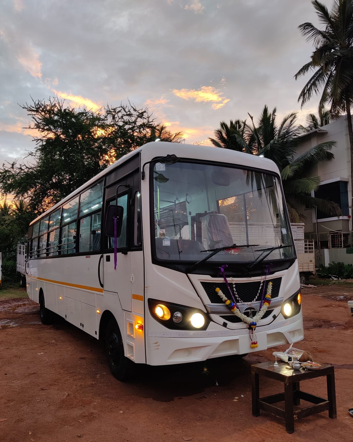 New white Tata bus with garland and headlights on, parked on red earth at sunset beside a small pooja table.