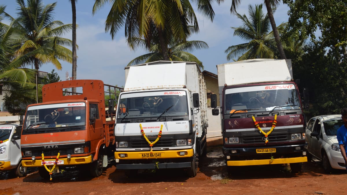 Three garlanded Tata trucks with Sushant windscreen decals parked under coconut palms against a blue sky.