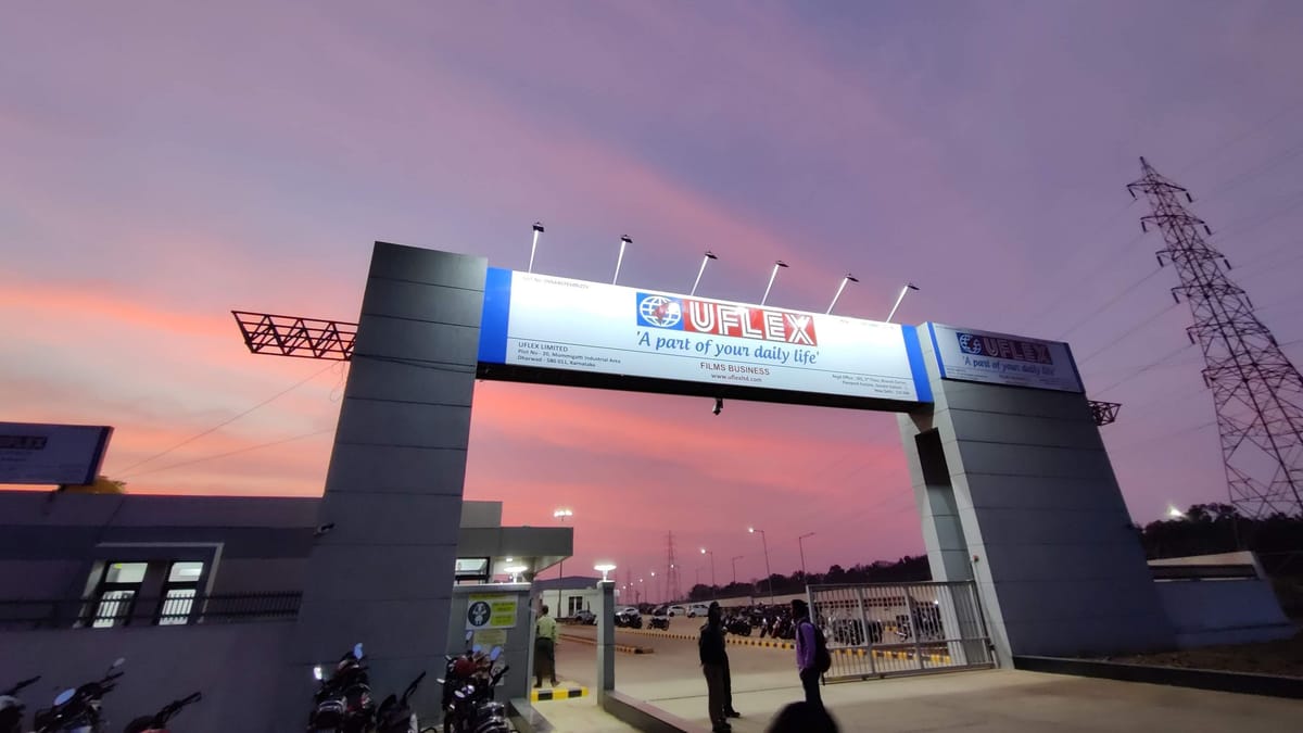 Illuminated overhead gate arch sign reading UFLEX at a factory entrance, lit by floodlights against a pink dusk sky.