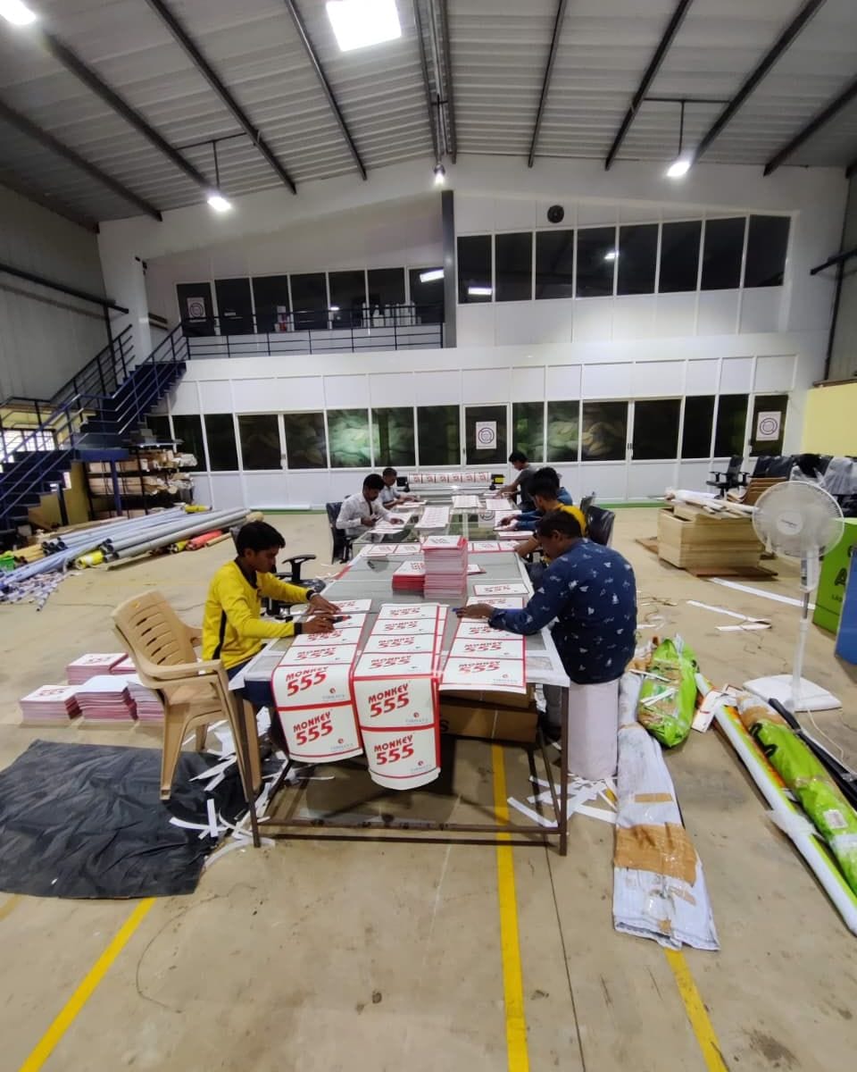 Four workers seated at long tables trimming stacks of printed white and red signage boards.
