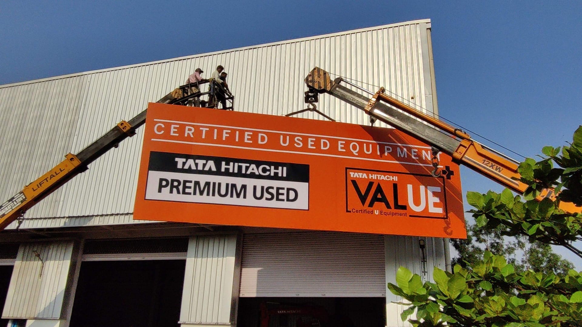 Crew on a crane fixing a Tata Hitachi dealership fascia to a showroom front